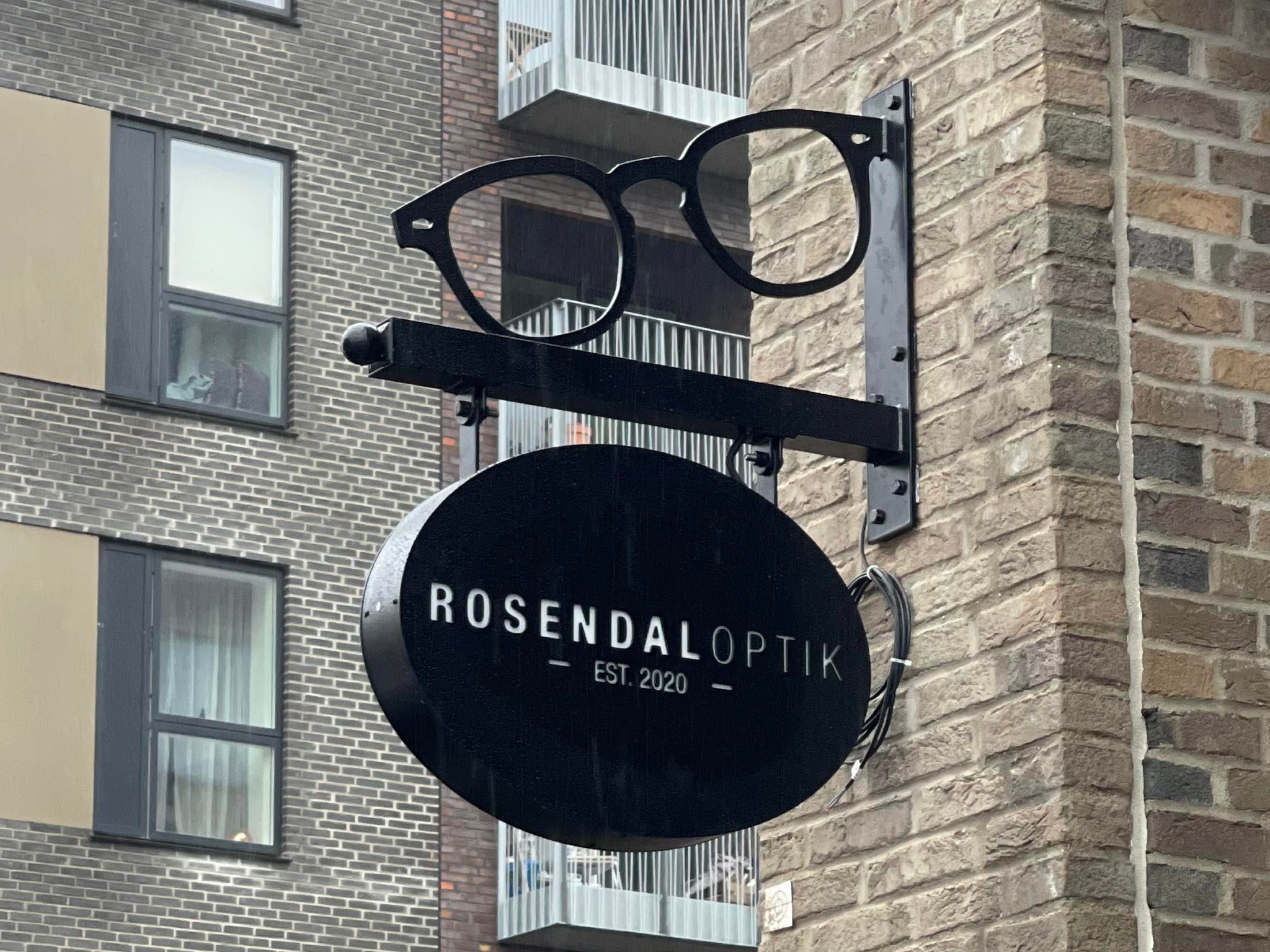 Sort udhængsskilt med briller og teksten 'Rosendal Optik' hængende på en mur.
