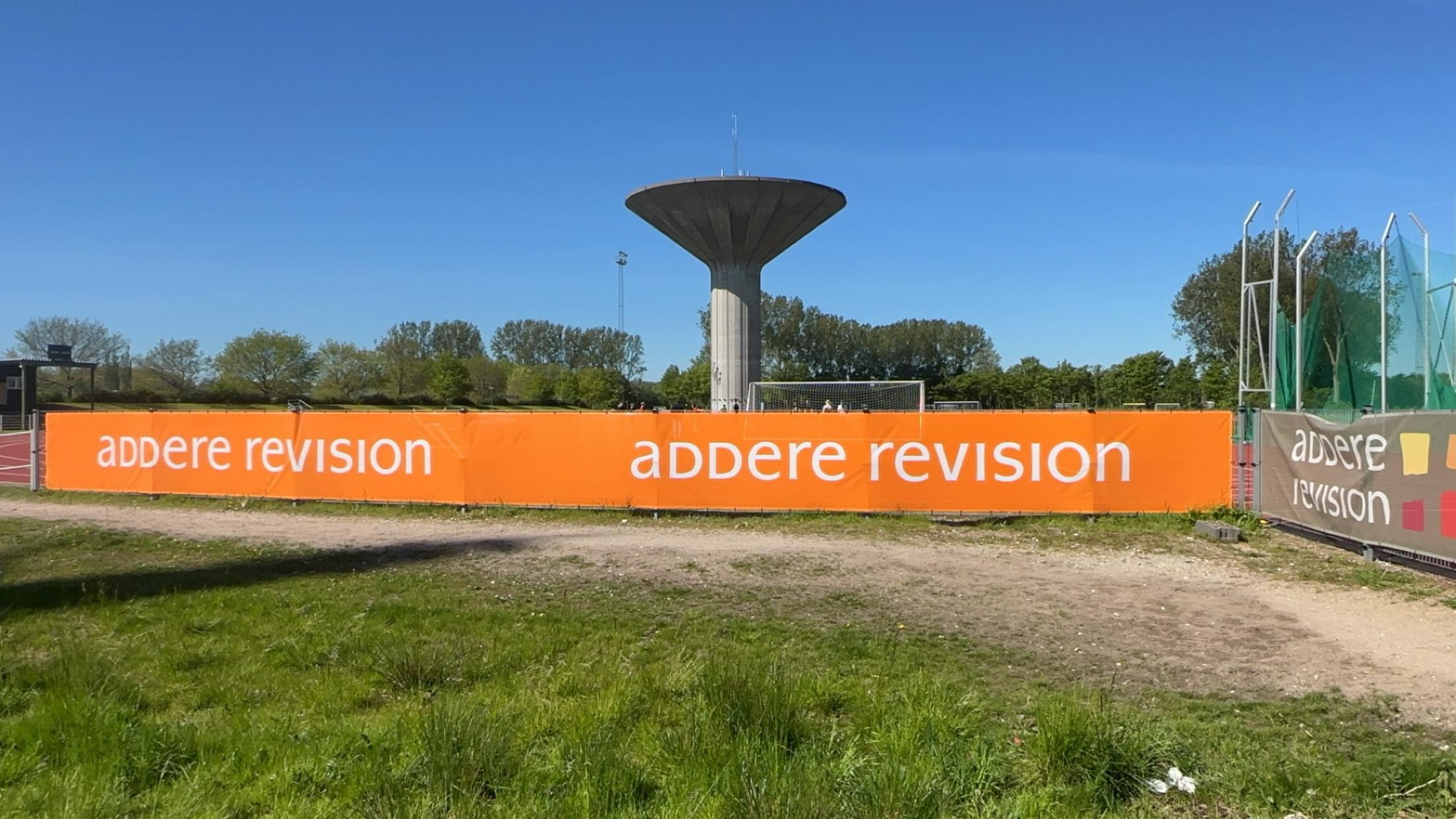 Banner med teksten 'addere revision' på en sportsplads, med en vandtårn i baggrunden.