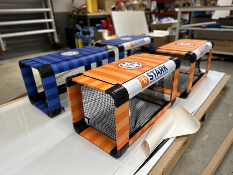 Tre farverige fodboldmål i blå, orange og stribet design fra Stark, placeret på en arbejdsflade.