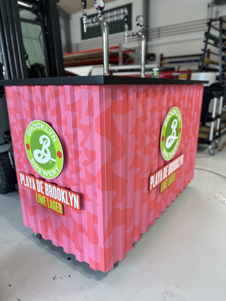 Bar med logo fra Brooklyn Brewery og teksten 'Playa de Brooklyn Lime Lager' på en farverig overflade.