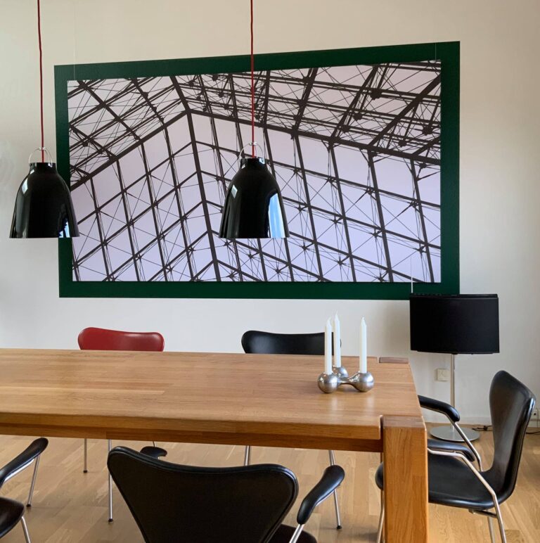 Stort akustikbillede med print af en metalstruktur hængende over et træbord med sorte stole og lamper.