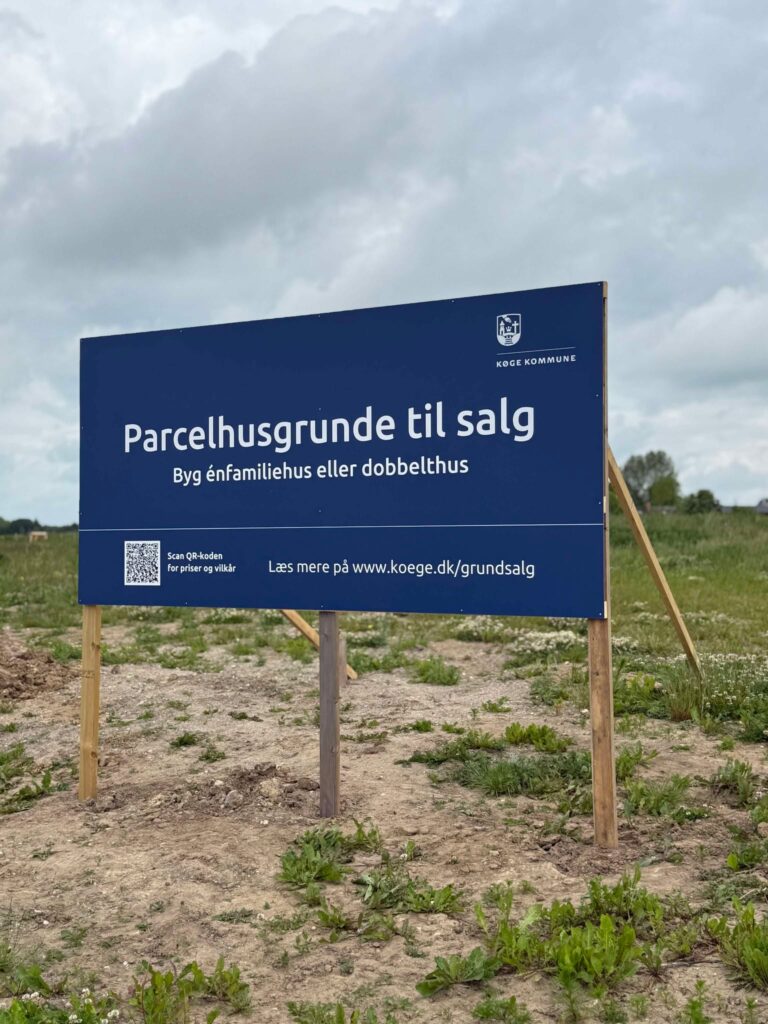 Blå skilt fra Køge Kommune med teksten 'Parcelhusgrunde til salg' på en byggeplads.