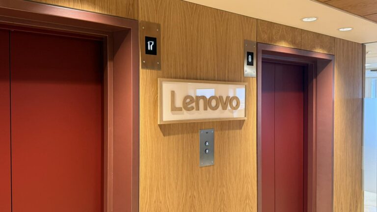 Indendørs elevatorområde med træpaneler og to elevatordøre; centralt Lenovo-signet på trævæggen, glasdør i baggrunden og loftsbelysning.