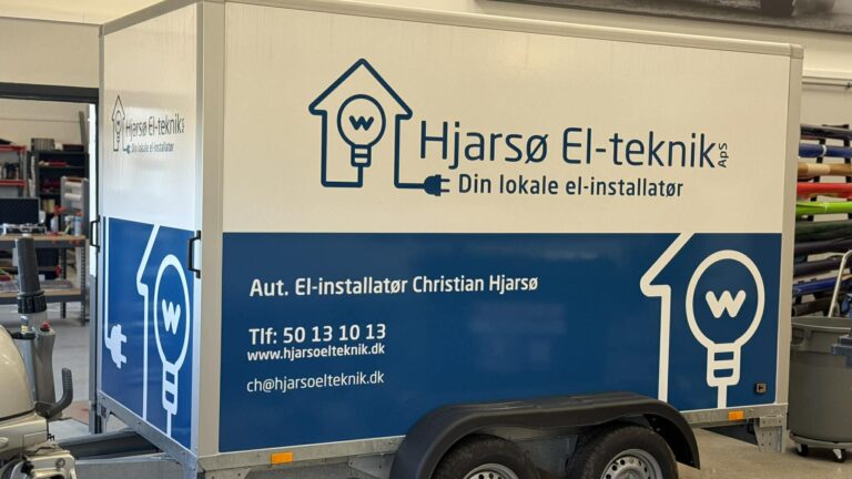 Blå og hvid trailer med branding for Hjarsø El-teknik, aut. el-installatør Christian Hjarsø, visende logo og kontaktoplysninger i værkstedsmiljøet