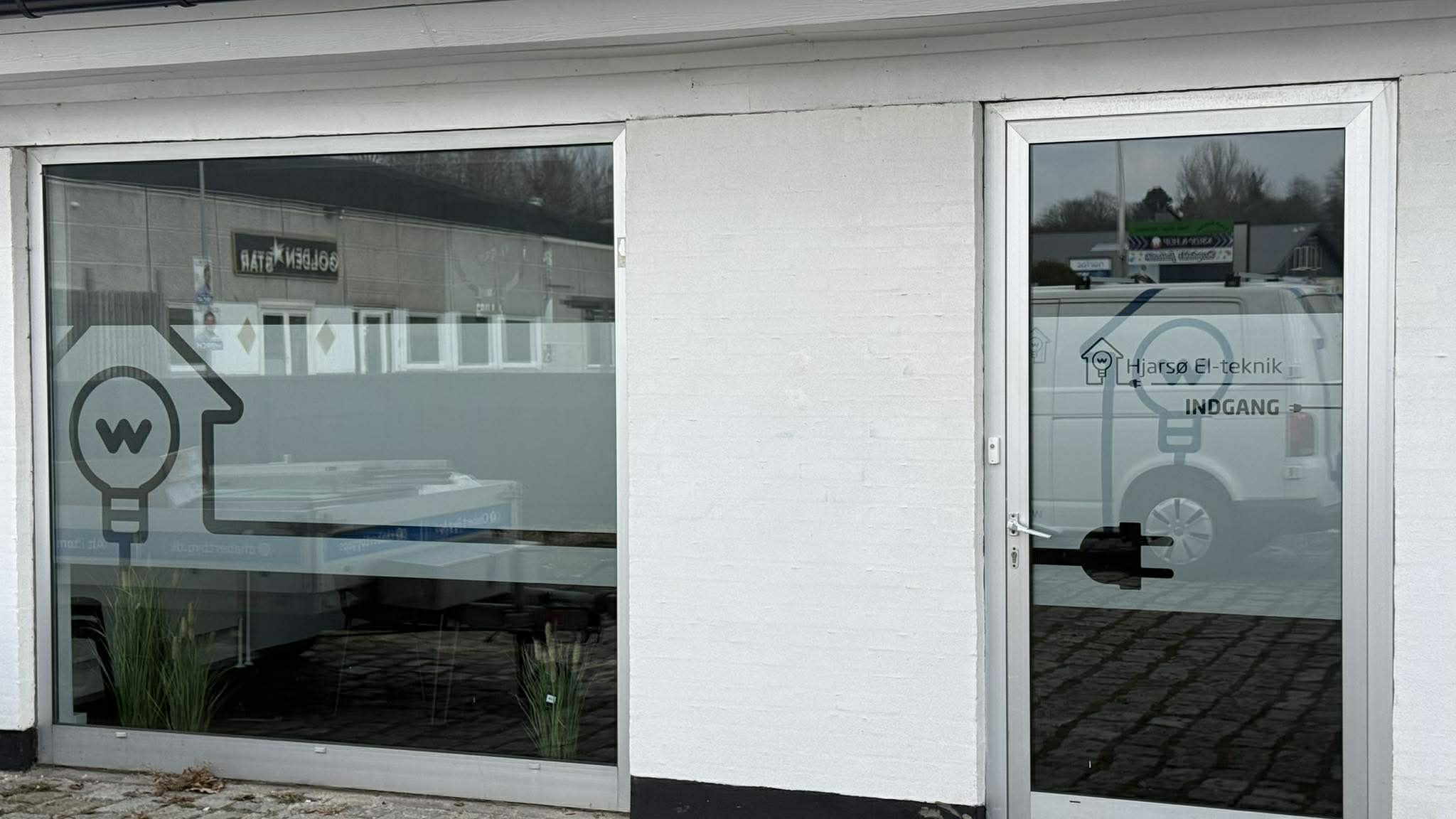 Glasfacaden ved Hjarse El-teknik har mat folie, logo på ruden og teksten INDGANG på døren foran den hvide mur.