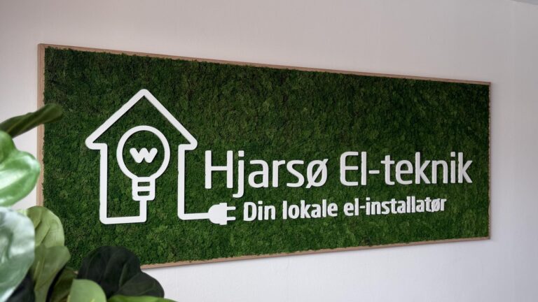 Det grønne vægskilt viser Hjarsø El-teknik med logo og teksten "Din lokale el-installatør" på en lys væg.