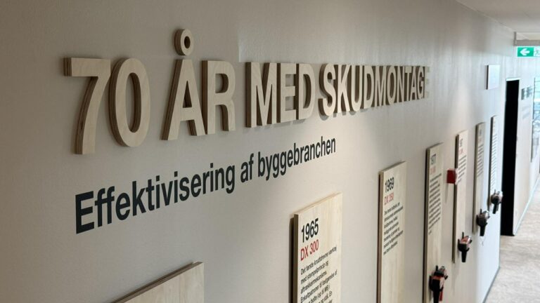 I gangen står 70 ÅR MED SKODMONTAGE sammen med Effektivisering af byggebranchen og historiske plaketter på en lys væg i en lang korridor.