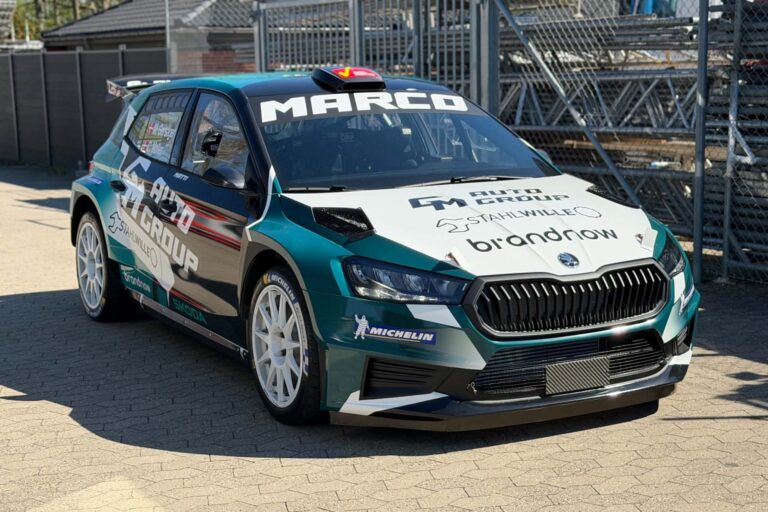 Med Brandnow- og MARCO-dekoration står en Škoda Fabia RS Rally2 med Michelin-dæk og turkis-hvid lakering foran et industrihegn.