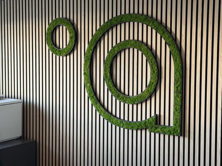 På en lodret trælamelvæg ses MOS-logoet udført i grøn mos og sort ramme, som giver en markant, naturinspireret vægdekoration.